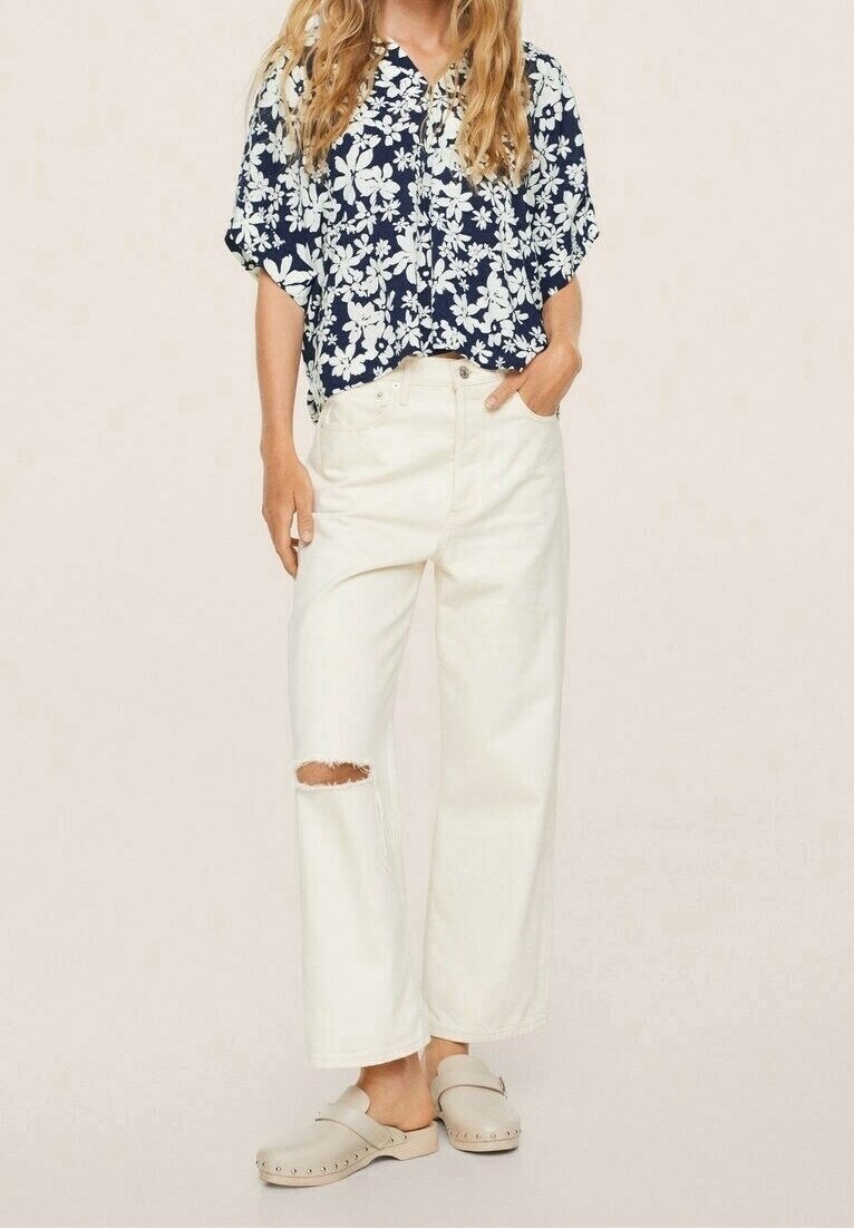 Femme portant un chemisier bleu marine à fleurs, un pantalon blanc taille haute à jambes larges avec une déchirure au genou, et des sabots beige à enfiler.