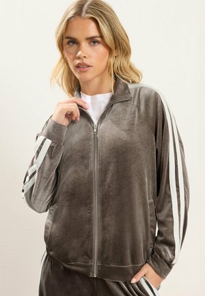 Lyshåret kvinde iført en grå velour træningsjakke med hvide striber på ærmerne, holder lynlåsen nær kraven, stående mod en ensfarvet baggrund.