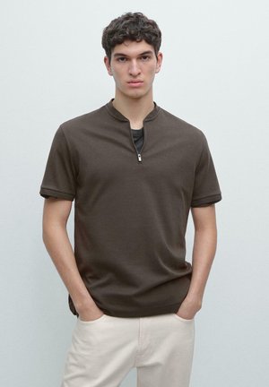 Jonge man met kort krullend haar, gekleed in een donkerbruin shirt met korte mouwen en een ritskraag, en lichtbeige broek, handen in de zakken, neutrale achtergrond.