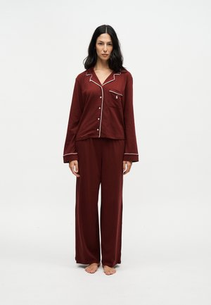 Woman standing barefoot wearing a dark red long-sleeve buttoned pajama set with white trim on collar, cuffs, and pocket.