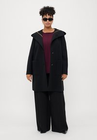 Cappotto di lana nero con cappuccio, due tasche frontali e dettagli di cucitura arancioni. Indossato sopra un top borgogna e pantaloni a gamba larga neri.