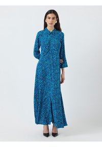 Robe à manches longues avec boutons, de couleur bleue et ornée d'un motif floral. Elle présente un ourlet évasé et un tissu léger pour plus de confort. Portée avec des escarpins noirs.