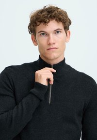 Schwarzer Stehkragenpullover aus strukturiertem Strickstoff, mit einem hohen Kragen und gerippten Bündchen, Model hält den Reißverschluss-Zipper.