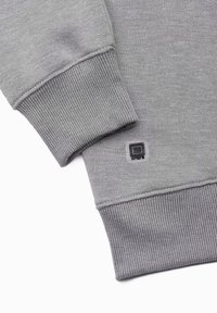 Manica e polsino di una felpa grigia con texture a coste, che mostrano un piccolo logo quadrato nero e grigio vicino all'orlo del polsino.
