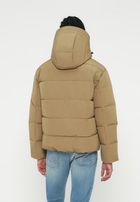 Khaki pufferjakke med hætte, med quiltede sektioner og en afslappet pasform. Parret med blå jeans, der viser en glat stoftekstur.