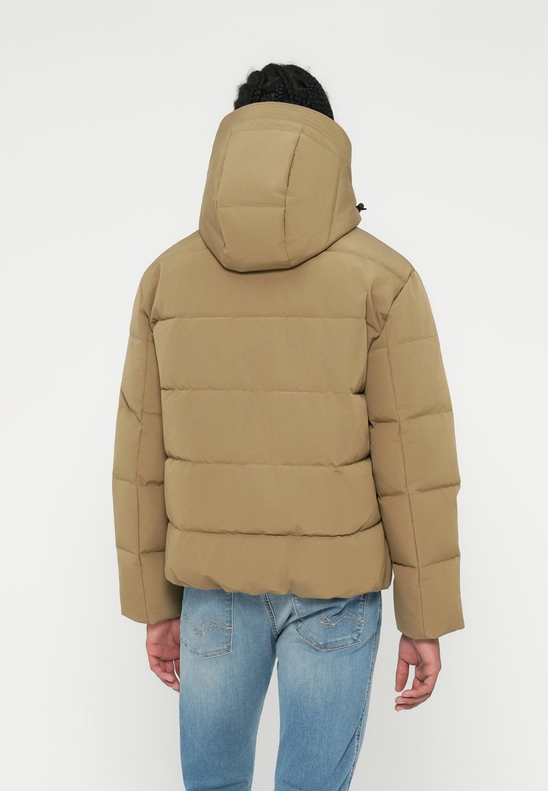 Khakifarbene Steppjacke mit Kapuze, die Steppnähte und eine lockere Passform aufweist. Kombiniert mit blauen Jeans, die eine glatte Stoffoberfläche zeigen.