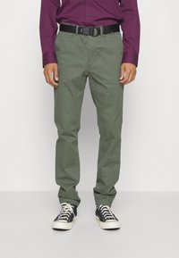 Pantaloni di cotone verdi con un design a gamba dritta, abbinati a una cintura nera regolabile e sneakers bianco e nere. Tessuto di trama strutturata.