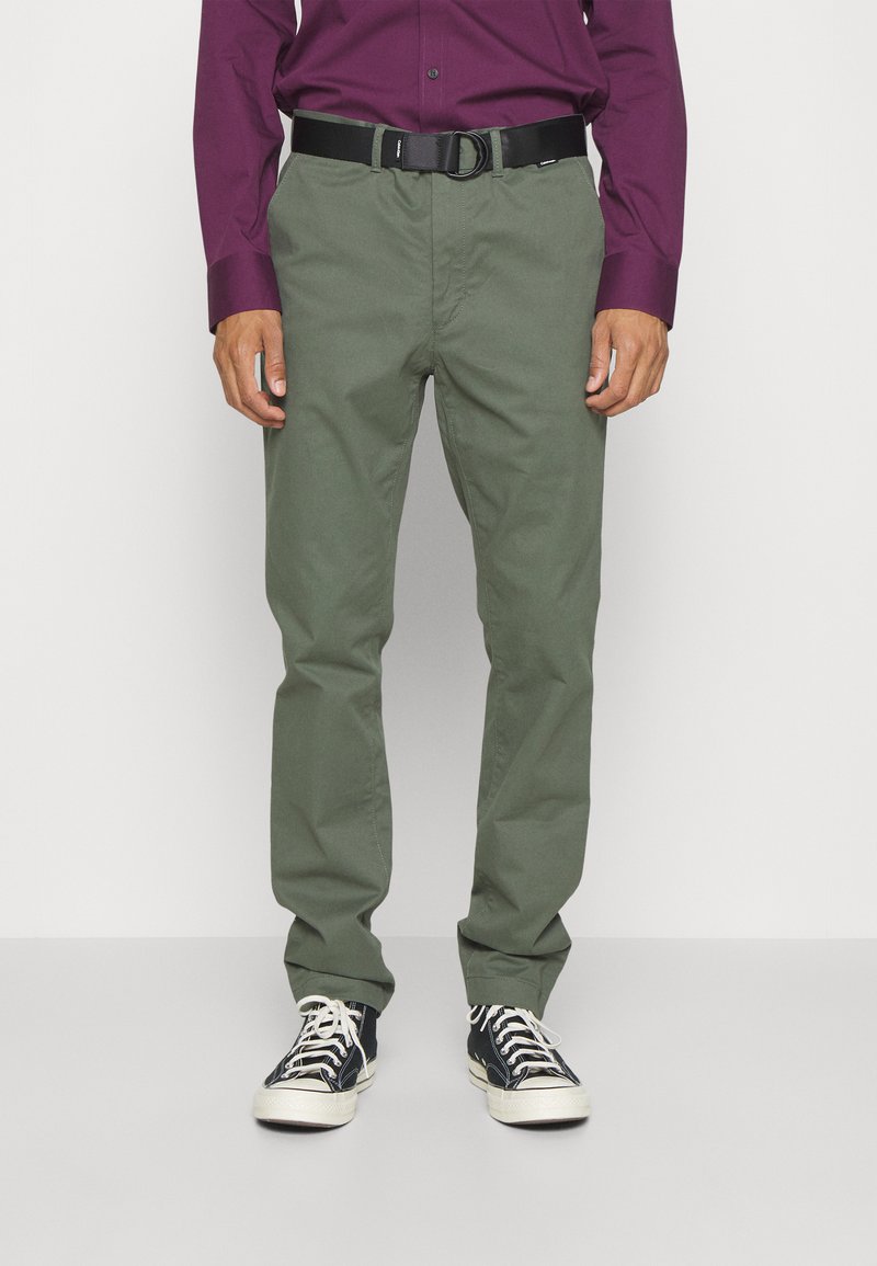 Pantaloni di cotone verdi con un design a gamba dritta, abbinati a una cintura nera regolabile e sneakers bianco e nere. Tessuto di trama strutturata.