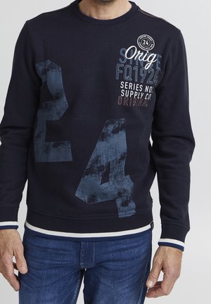Homme portant un sweat-shirt bleu marine avec un grand imprimé « E4 » délavé et le texte « Orig FQ1924 Series No Supply Co. Original » sur le devant, associé à un jean bleu.