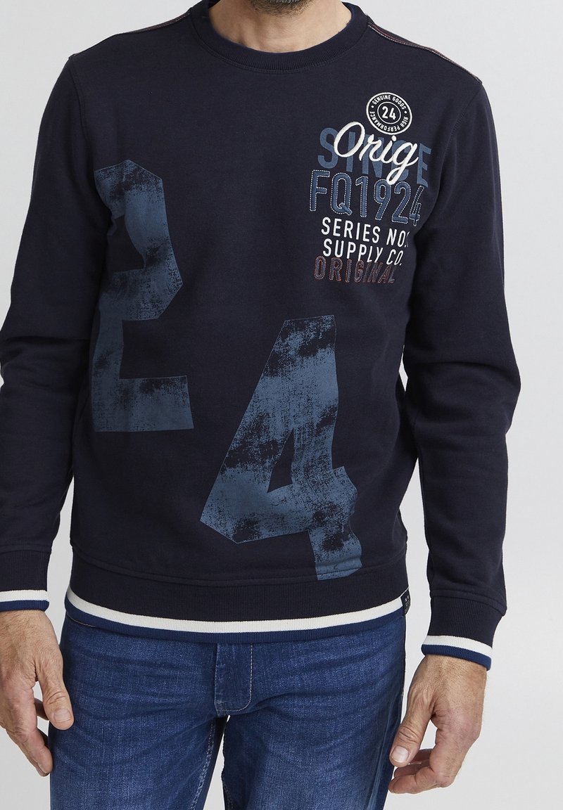 Homme portant un sweat-shirt bleu marine avec un grand imprimé « E4 » délavé et le texte « Orig FQ1924 Series No Supply Co. Original » sur le devant, associé à un jean bleu.
