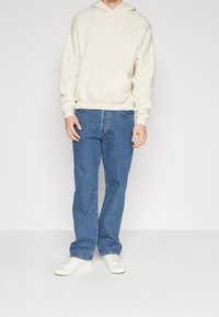 Beige huvtröja med en framficka, i kombination med blå raka jeans och vita sneakers. Minimalistisk design, avslappnad passform.