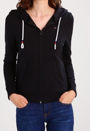 Haut de survêtement noir à zip fabriqué en matériau doux, avec une capuche, des cordons de serrage blancs avec des accents colorés, et des poches avant.