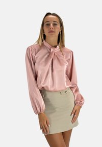 Hellrosa Satinbluse mit langen, voluminösen Ärmeln und geknotetem Kragen, kombiniert mit einem beigefarbenen, figurbetonten Minirock mit Seitentaschen und Knopf.