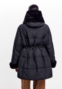Sort polstret frakke med en indsnævret talje, faux fur krave og manchetter samt quiltet design. Bagview med hætte og langtærs efter.