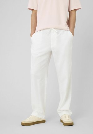 Personne portant un pantalon blanc ample à cordon, un t-shirt rose pâle à manches courtes et des baskets beiges à semelles en gomme, mains dans les poches.