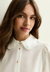Jeune femme aux longs cheveux bruns portant une blouse blanche avec un col plissé et des boutons dorés, regardant légèrement souriante vers la caméra.
