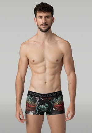 Atletický muž s krátkymi tmavými vlasmi a bradou, oblečený v čiernych boxerkách s farebnými grafickými potlačami a pásom s logom značky.