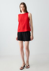 Donna in piedi che indossa un top rosso senza maniche, pantaloni corti neri e sandali col tacco neri su uno sfondo chiaro e semplice.