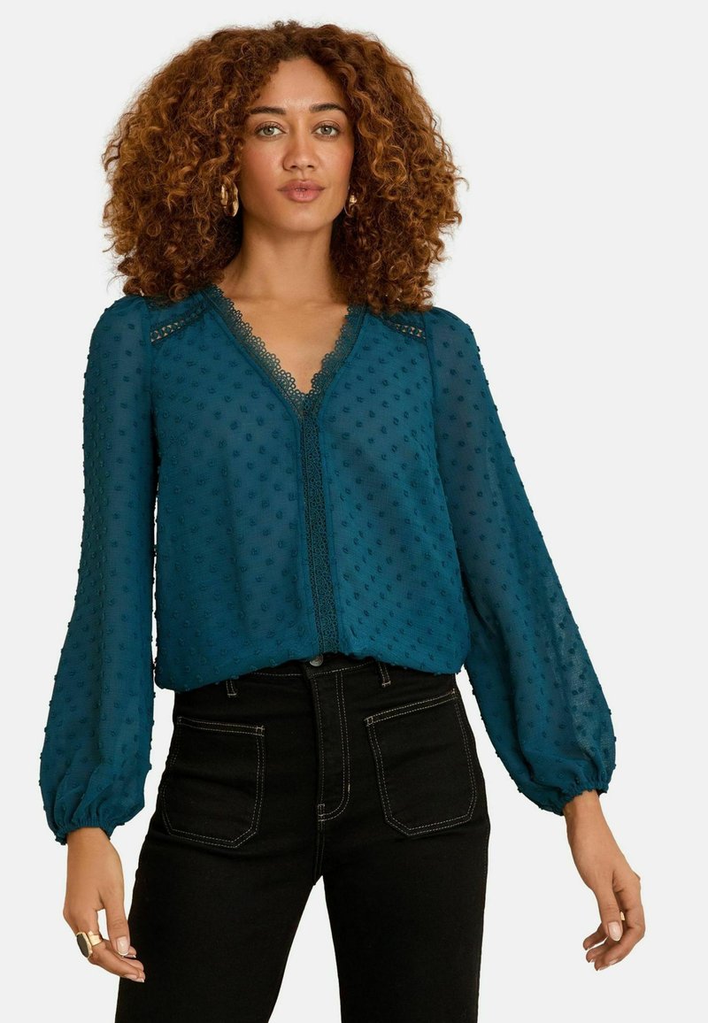 Tīrkīza, caurspīdīga blūze ar tekstūrētiem punktiem, garām pūkainām piedurknēm un mežģīņu apdari gar V veida kaklu. Pāris ar augsto jostasvietu melnām džinsām.