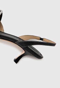 Fekete bőr macskasarkú szandál bézs bokapánttal és a talpon, valamint a pánton látható fehér varrással, fehér háttér előtt.