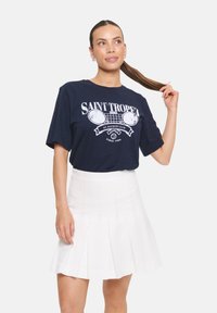 T-shirt bleu marine avec impression graphique blanche représentant des raquettes de tennis et du texte, associé à une jupe plissée blanche.