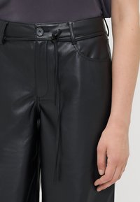 Pantalons en cuir noir avec une texture lisse, dotés d'une fermeture à bouton, d'un cordon de serrage ajustable et de deux poches latérales.