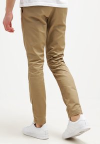 Pantaloni slim-fit color khaki con una texture liscia, caratterizzati da una gamba sfilata, abbinati a sneakers bianche da sport.