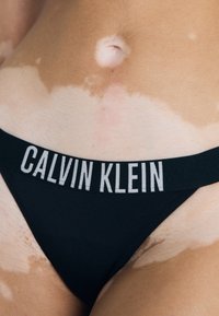 Czarny thong Calvin Klein z szerokim pasem, na którym znajduje się nazwa marki w białym napisie, prezentujący gładką teksturę materiału i minimalistyczny design.