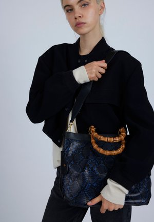 Giacca nera corta con accenti beige; il modello tiene una borsa blu in pelle di serpente con manico in bambù e dettagli dorati, su uno sfondo chiaro.