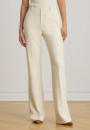 Kvinde iført cremefarvede, vidde ben bukser med højtaljet snit, der lukkes med knap og lynlås, kombineret med brune spidse sko på trægulv.