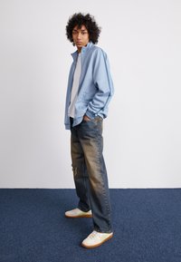 Világoskék sportdzseki állógallérral, amelyet koptatott kék farmernadrággal és bézs sportcipővel viselnek, zöld részletekkel.