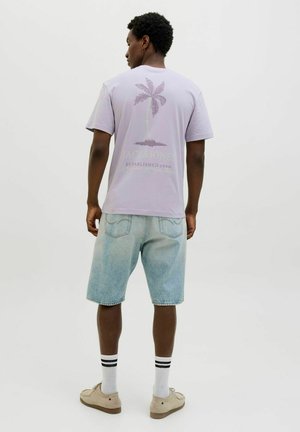 Helles lila T-Shirt mit einem Palmen-Design, kombiniert mit hellblauen Jeansshorts und beigen Schuhen. Sichtbare schwarz-weiße gestreifte Socken.
