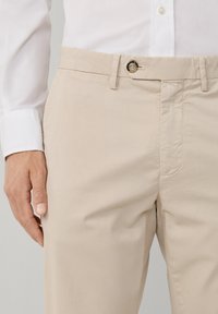 Chino in cotone beige con taglio dritto, chiusura con bottone e tasche laterali, esposti su uno sfondo chiaro.