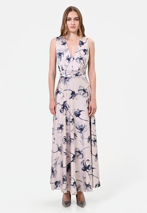 Femme debout portant une robe maxi sans manches rose clair avec un imprimé floral foncé et un profond décolleté en V, associée à des chaussures grises pointues.