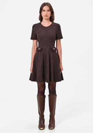 Donna che indossa un vestito marrone a maniche corte con tasche con bottoni, collant neri e stivali di pelle marrone fino al ginocchio, in piedi dritta.