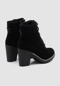 Botas negras hasta el tobillo con tacones gruesos, parte delantera con cordones, cuellos acolchados y suelas texturizadas, mostradas desde el lado y la parte posterior sobre un fondo liso.