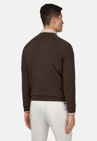 Homme portant un pull à manches longues marron foncé et un pantalon blanc, debout de dos sur un fond clair uni.