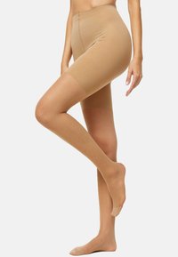 Frau in nahtlosen, hautfarbenen, hochgeschnittenen Shapewear-Shorts und transparenten Strumpfhosen, die mit einem leicht angehobenen Bein steht.
