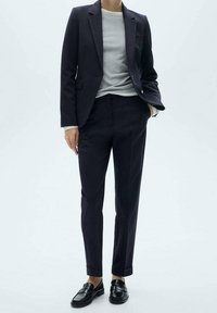 Námořnický oblek na míru se slim fit střihem, jednořadým sakem a kalhotami s plochým předním dílem. V kombinaci se světle šedým svetrem s kulatým výstřihem a černými polobotkami.