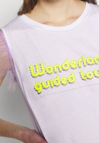 Světle purpurový bezrukávový top s žlutým nápisem "Prohlídka říše divů", s průhlednými levandulovými tylovými akcenty na ramenech. Měkká tkanina.