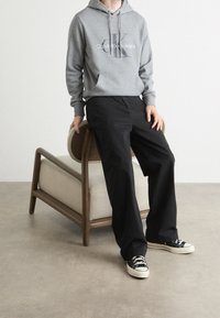 Persona con una sudadera gris de Calvin Klein, pantalones anchos negros y zapatillas negras apoyada en una silla de madera con cojín beige contra una pared blanca.