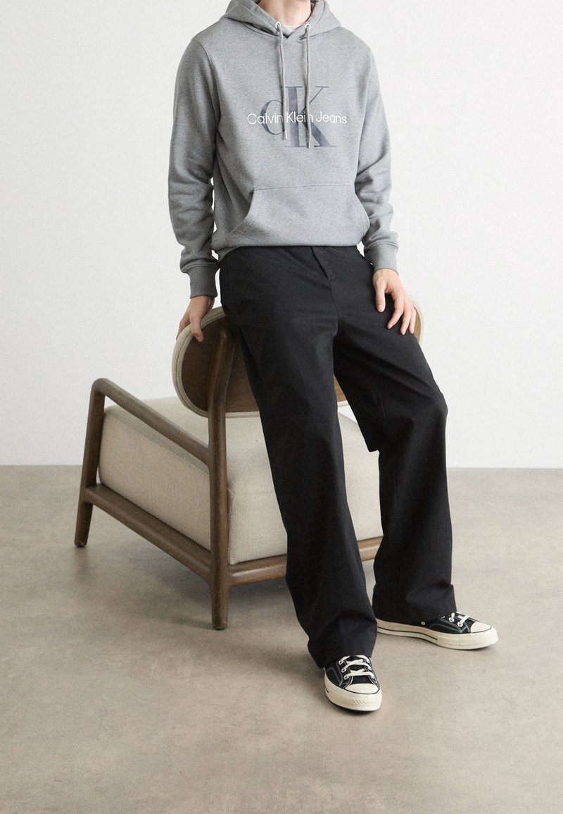 Persona con una sudadera gris de Calvin Klein, pantalones anchos negros y zapatillas negras apoyada en una silla de madera con cojín beige contra una pared blanca.