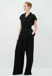 Svart skräddarsydd väst med bälte, korta ärmar, dubbelknäppt design, ihopparad med vida svarta byxor, slät tygtextur.