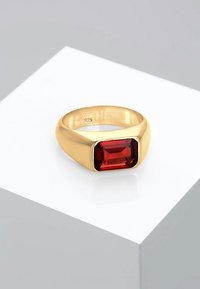 Goldfarbener Ring mit einem rechteckigen roten Stein, glatte Oberfläche, glatter Band, innen mit "925" markiert. Vor einem weißen Hintergrund.