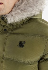 Olivengrøn pufferjakke med en foring af pels i hætten, ribbet tekstur, tryklås og et sort logo-patch på den øverste venstre side.