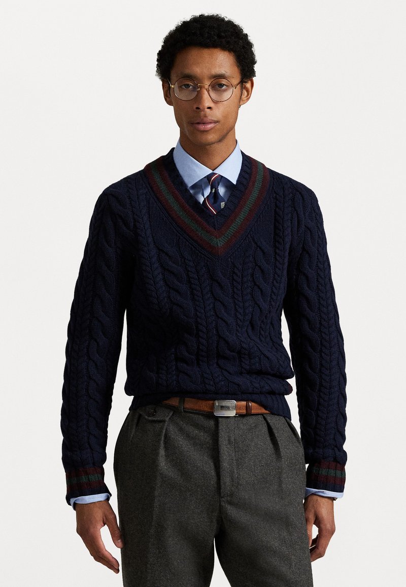 Maglione navy a trecce con scollatura a V e colletto a righe bordeaux-verde, indossato sopra una camicia col collo azzurra e pantaloni grigi.