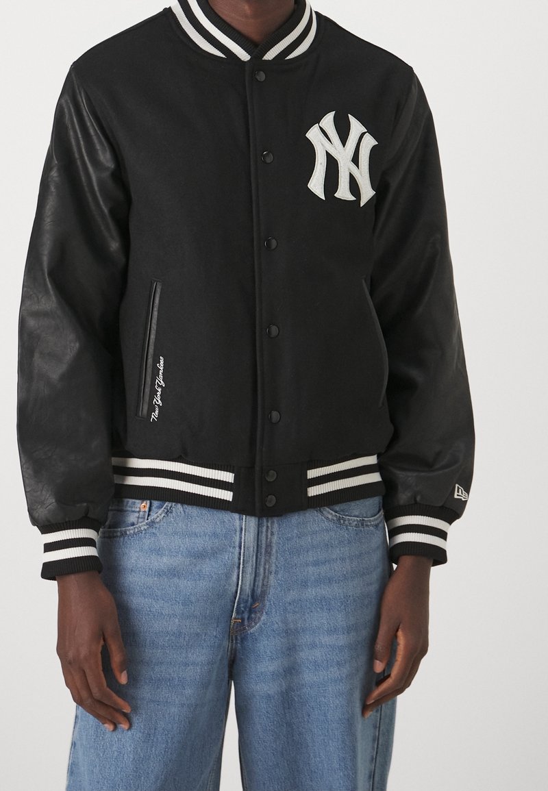 Svart varsityjacka med vita randiga manschetter och krage, New York Yankees-logotyp på bröstet, ihopparad med ljusblå jeans.