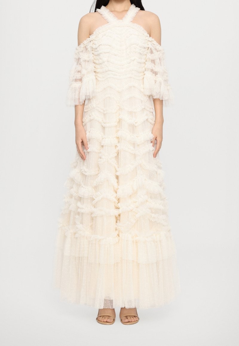 Femme portant une longue robe blanche épaules dénudées avec des volants et un tissu transparent, debout les bras détendus le long du corps.