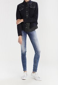 Mörkblå denimjacka med nitar på axlarna, tillsammans med en mörk skjorta, ljusblå skinny jeans och vita sneakers.