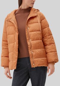Kobieta w pomarańczowej kurtce puchowej z rozpiętym zamkiem błyskawicznym, nosząca brązowy sweter z golfem i ciemne spodnie, stojąca na jednolitym tle.